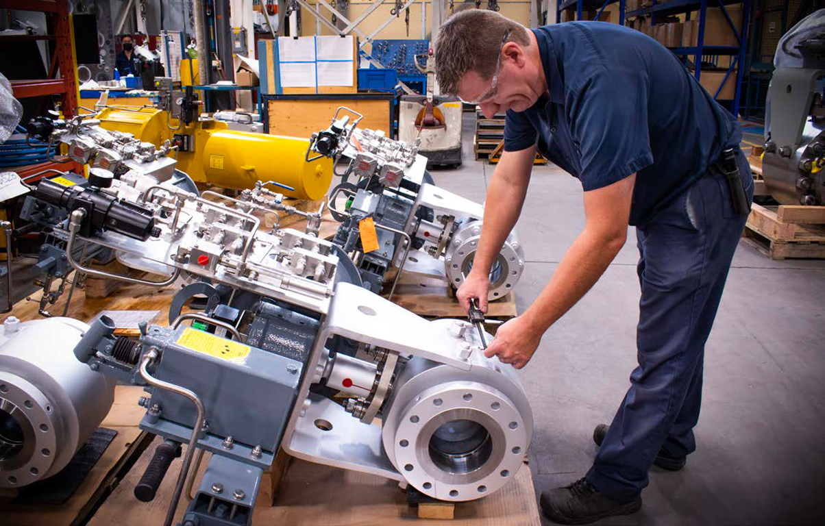 A man wearing safety glasses and a dark blue uniform is working on a large industrial machine in a factory setting. He is adjusting components with a wrench. Various tools and parts are visible around him in the well-organized workspace.