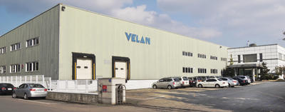 Un grand bâtiment industriel avec l'inscription « VELAN » sur le côté, avec des quais de chargement et une rangée de voitures garées. Le ciel est partiellement nuageux et la zone est dégagée, ce qui suggère une entreprise ou une usine.
