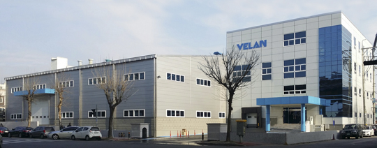 Image d'un complexe de bâtiments industriels avec deux structures reliées. Le bâtiment de gauche a une façade en métal, tandis que le bâtiment de droite, plus haut, a une conception en verre et en béton. Le nom « VELAN » est affiché sur la structure la plus haute. Plusieurs voitures sont garées à l'extérieur.