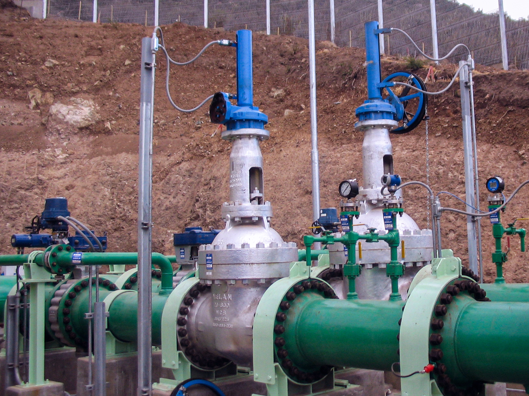 Two large industrial gate valves with blue actuators mounted on green pipelines outdoors, surrounded by gauges and control equipment, with a dirt embankment and fence in the background.