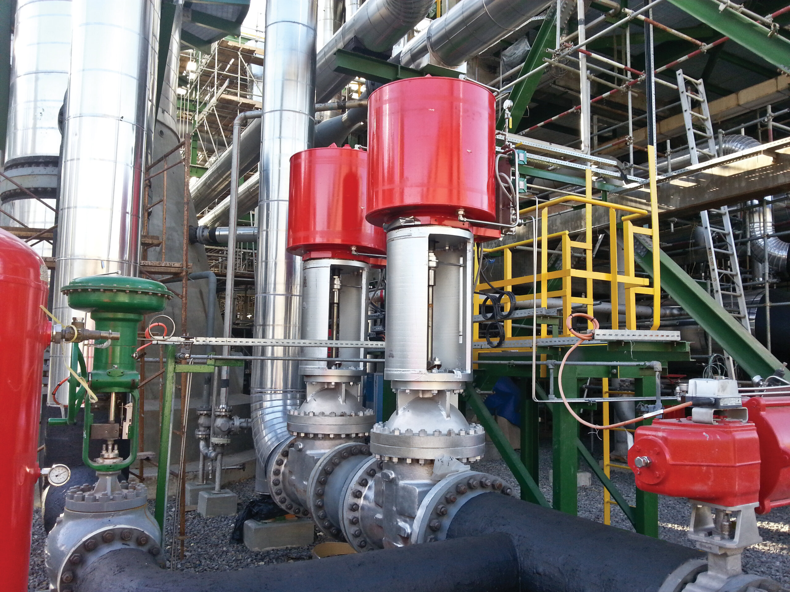 Large industrial valves with red actuators are installed on metal pipes at a processing plant, surrounded by scaffolding, yellow stairs, and other machinery. The scene appears outdoors with visible sunlight.