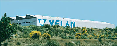 Un grand bâtiment industriel avec une façade blanche et l'inscription bleue "VELAN" est situé sur une colline verdoyante avec des buissons et des arbres au premier plan. Le ciel est clair et bleu.