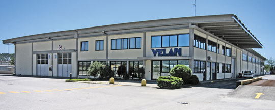 Un grand bâtiment industriel avec une façade beige et de nombreuses fenêtres. Le nom « VELAN » est affiché bien en évidence sur le côté. Le bâtiment est entouré d'une zone pavée et d'un peu de verdure sous un ciel bleu clair.
