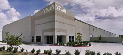 Un grand bâtiment industriel avec le nom « VELAN » sur la façade. Il a une façade beige, quelques arbres et arbustes autour et une allée pavée sous un ciel partiellement nuageux.