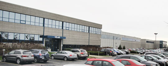 Un long immeuble de bureaux de plain-pied avec de grandes fenêtres et une enseigne à l'entrée. Le parking en face est rempli de plusieurs voitures garées. Le ciel est couvert.