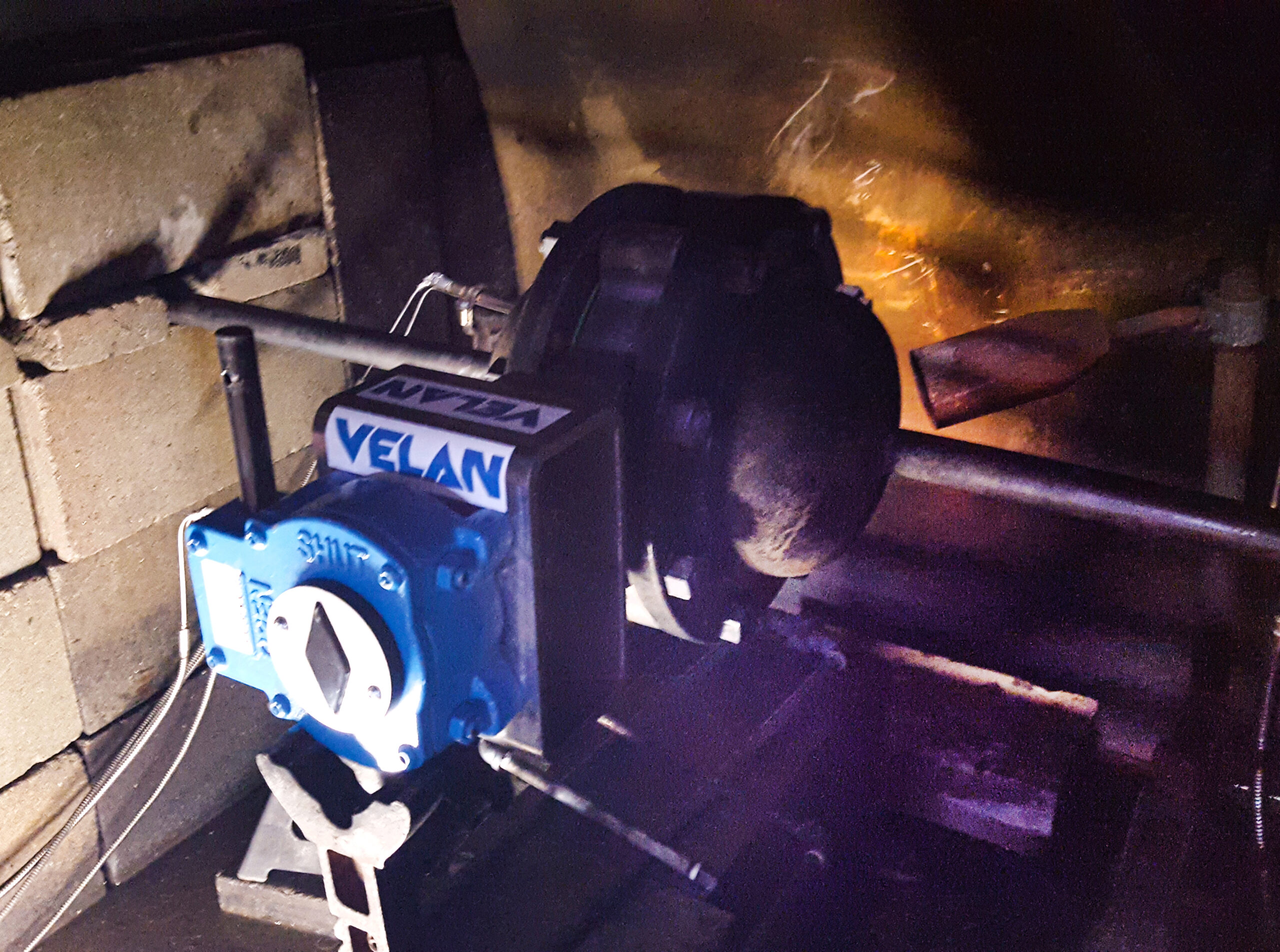 Une vanne industrielle bleue en Velan fixée à un système de tuyaux métalliques, avec un mur de briques en arrière-plan et une grande roue ou un raccord rouillé derrière la vanne. La zone est faiblement éclairée, avec une lumière orange sur le côté droit.
