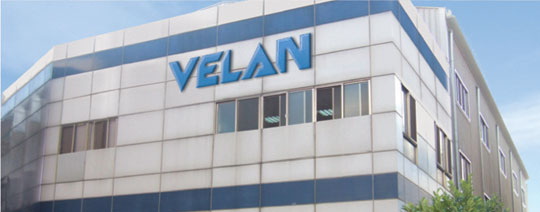Un grand bâtiment dont la façade est faite de panneaux de verre et de métal, affichant le mot « VELAN » en lettres bleues. Plusieurs fenêtres reflétant un ciel dégagé sont visibles, et de la verdure est présente dans le coin inférieur droit.