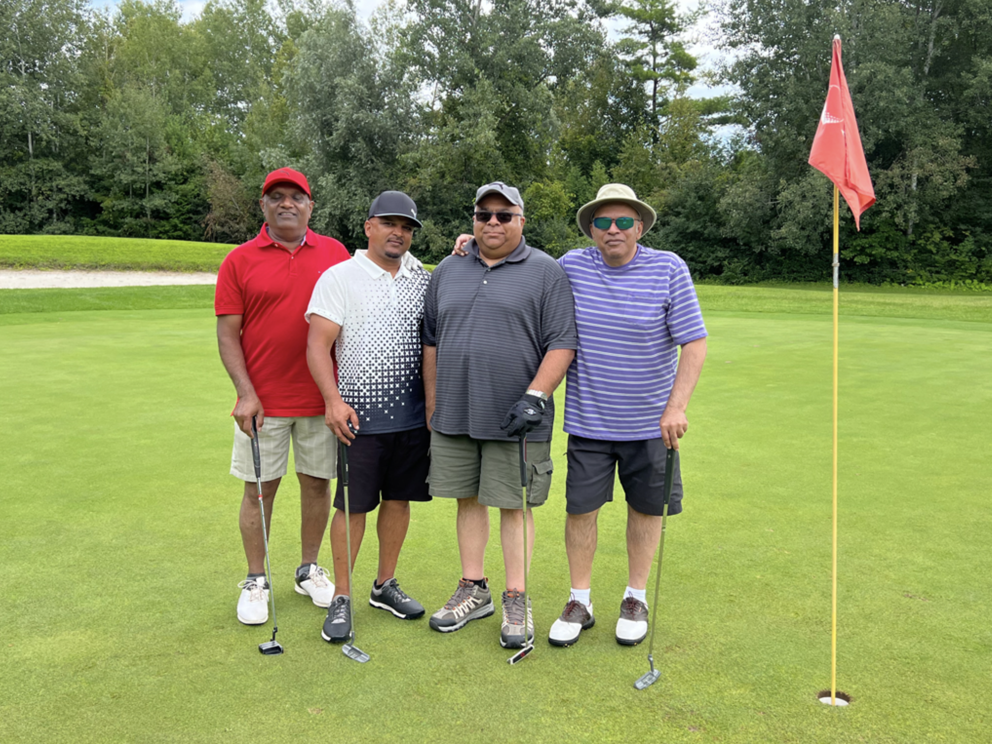 Four men standing on a golf course green with putters, posing by a hole marked with a red flag. They are dressed in casual golf attire including hats and sunglasses. Trees and shrubs are in the background.