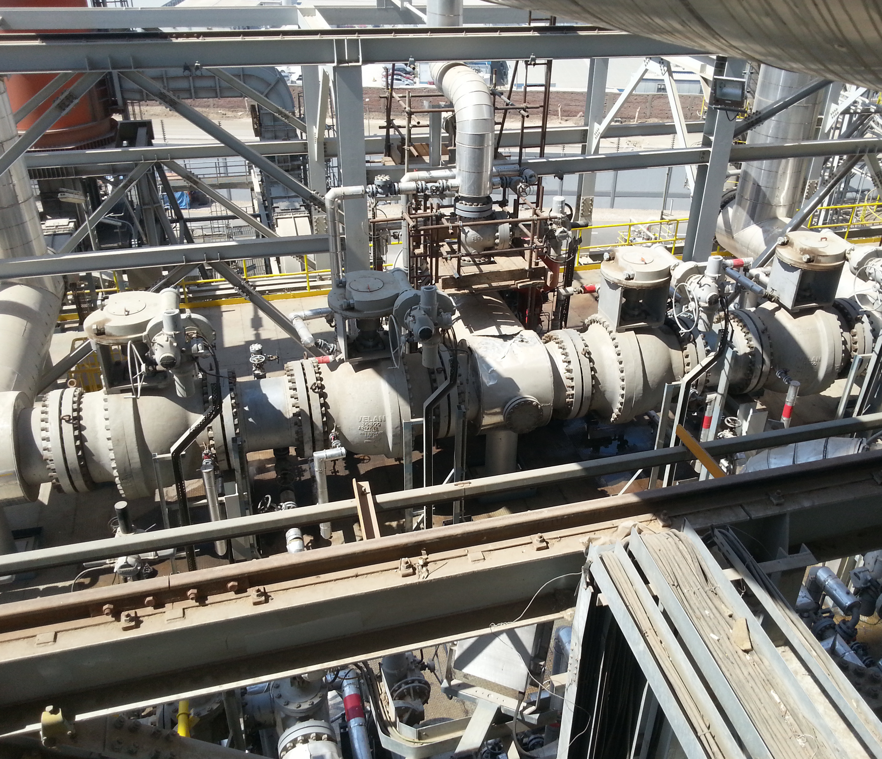 Une grande installation industrielle avec des tuyaux métalliques et des machines interconnectées.Des appareils de robinetterie et des jauges sont visibles, avec des structures métalliques substantielles et plusieurs niveaux.L'arrière-plan montre des équipements industriels supplémentaires et un aperçu de l'environnement extérieur.