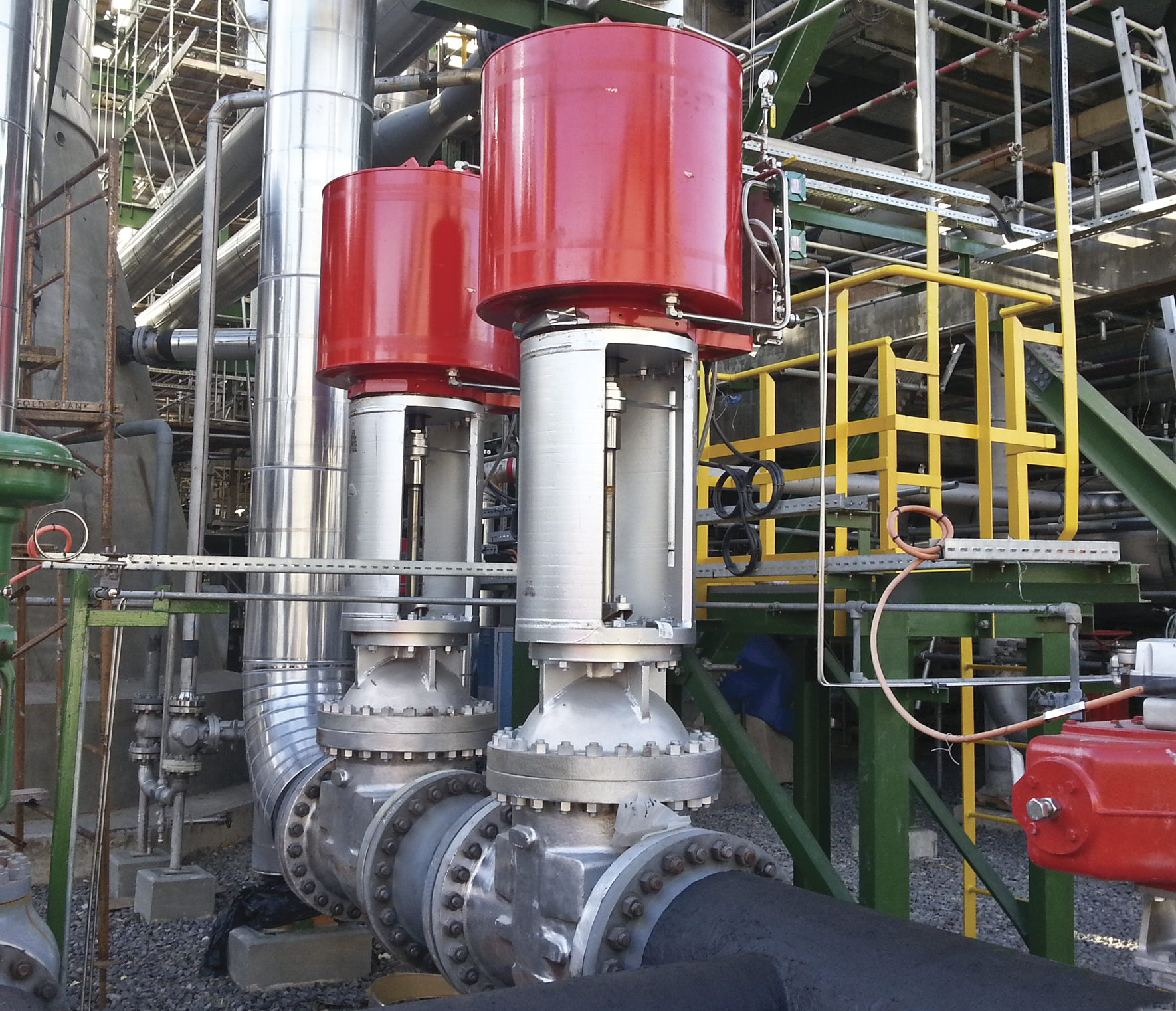 Deux grands appareils de robinetterie industriels avec actionneurs rouges installés sur une canalisation au sein d'une installation complexe. Le décor comprend divers tuyaux en acier, des structures de support vertes et des garde-corps de sécurité jaunes, suggérant un environnement industriel ou de fabrication.