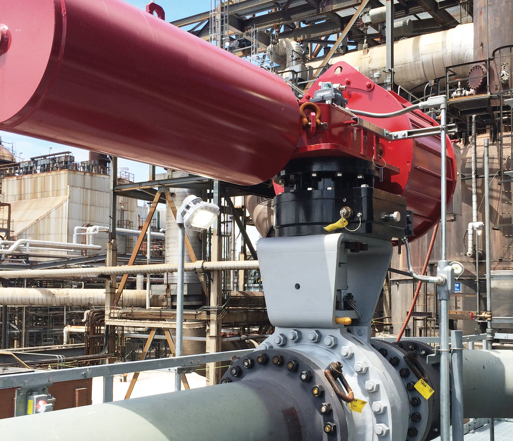Un environnement industriel comprenant un grand actionneur d’appareil de robinetterie relié à une canalisation. L'actionneur est rouge et repose sur un appareil de robinetterie gris avec de nombreux boulons. L'arrière-plan révèle un réseau complexe de tuyaux et de structures industrielles.