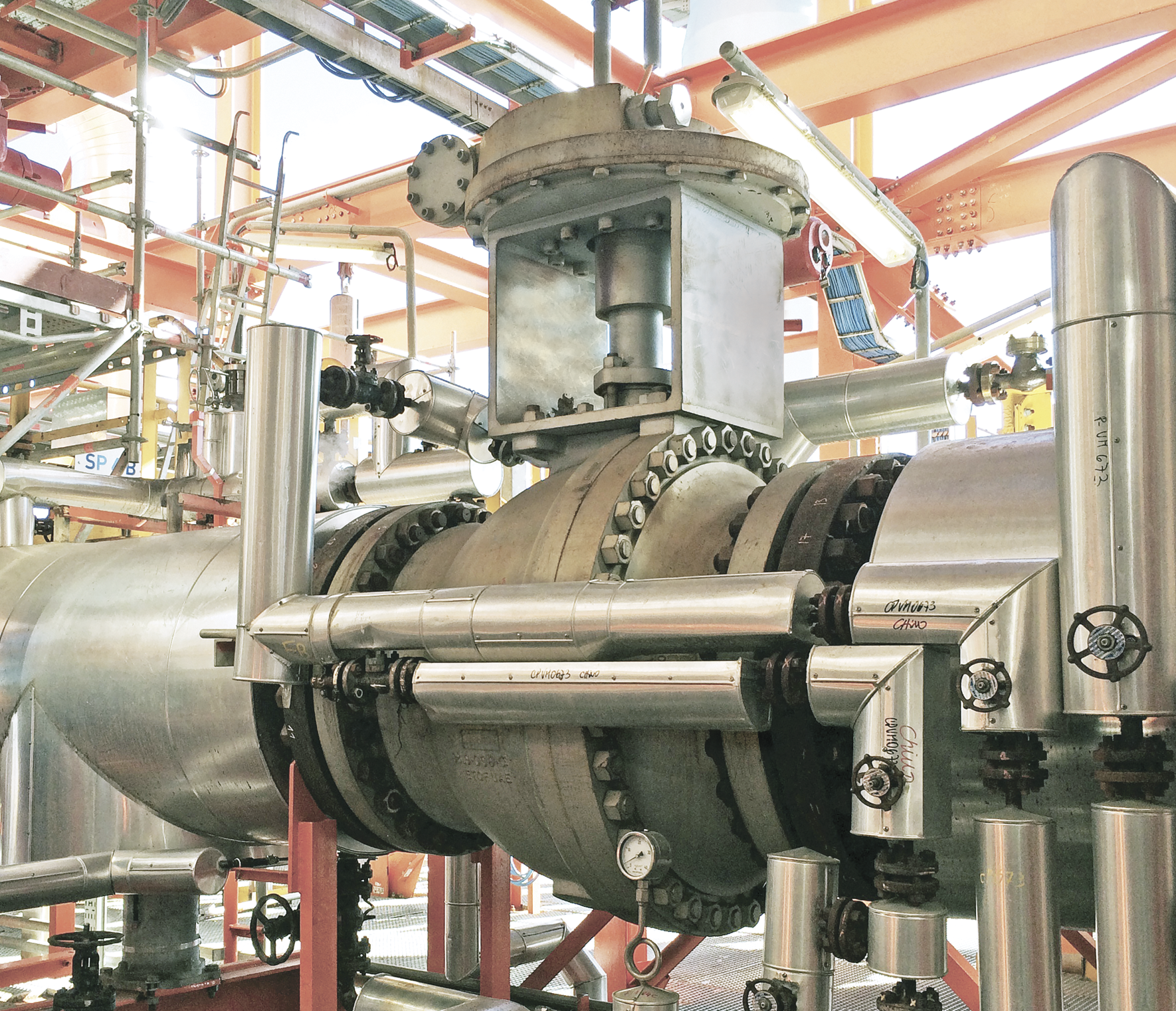 Vue rapprochée de la tuyauterie et de l’équipement industriels dans une usine de fabrication ou de transformation. L'installation comprend de gros tuyaux en acier, des appareils de robinetterie, des brides et divers raccords métalliques, avec une structure orange en arrière-plan.